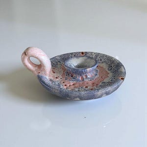 May include: A ceramic candle holder with a handle. The holder is a mix of colors, including pink, blue, and white. The candle holder has a circular base with a raised center for the candle. The handle is curved and has a hole.