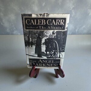 Op de afbeelding: Een hardcover boek getiteld "The Angel of Darkness" van Caleb Carr, auteur van "The Alienist". De omslag toont een zwart-wit afbeelding van een straatbeeld met figuren en een paardenkoets. Het boek wordt gepresenteerd op een kleine, rode houten standaard.