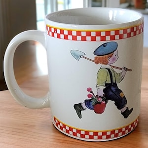 May include: White ceramic mug with a red and white checkered border. The mug features a cartoon illustration of a child carrying a shovel and a bucket of flowers.