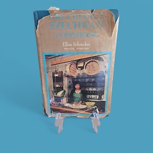 Puede incluir: Un libro de cocina vintage titulado "Mrs. Chiang's Szechwan Cookbook" de Ellen Schrecker. La portada muestra una foto de una mujer en una cocina preparando comida. El libro se exhibe sobre un soporte de plástico transparente sobre un fondo azul.