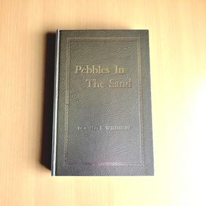Op de afbeelding: Een hardcover boek getiteld "Pebbles In The Sand" door Donald E. Wildmon. Het boek heeft een getextureerde, donkergrijze omslag met gouden letters en een decoratieve rand. Het boek ligt op een lichtbruin oppervlak.