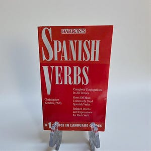 May include: A red book titled "SPANISH VERBS" by Barron's. The book includes complete conjugations, over 300 commonly used Spanish verbs, and related words. The author is Christopher Kendris, Ph.D. The book is displayed on a clear plastic stand.