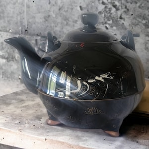 May include: A black teapot with gold accents and a floral design. The teapot has a rounded body and a spout on the side. The teapot is sitting on a grey surface.