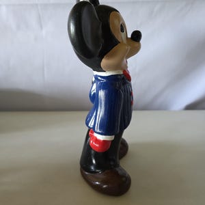 K&ouml;nnte beinhalten: Keramikfigur einer Zeichentrickmaus in einem blauen Anzug mit roten Akzenten, schwarzen Schuhen und roten Handschuhen. Die Maus hat gro&szlig;e schwarze Ohren, ein hellbraunes Gesicht und eine schwarze Nase. Die Figur steht aufrecht.
