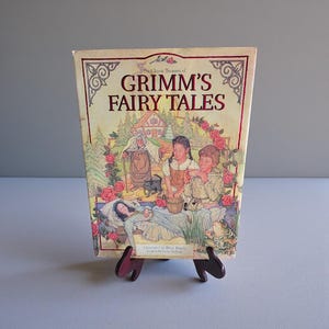 May include: A vintage copy of "Grimm's Fairy Tales" book, displayed on a wooden stand. The cover features a colorful illustration with the title in red lettering. The illustration depicts scenes from the fairy tales, including characters and settings.