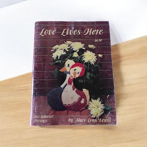 May include: A vintage book titled "Love Lives Here" with a brick-like background. The cover features a charming illustration of two ducks, one in a red dress and the other in a blue outfit, surrounded by white flowers. The book is by Mary Lynn Lewis.