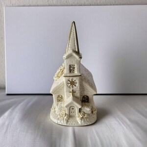 May include: A white ceramic church figurine with gold accents. The church has a steeple, a cross on the roof, and a window with a sun design. The figurine is decorated with floral details around the base.