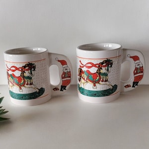 Puede incluir: Dos tazas de cerámica blanca con temática navideña. Cada taza presenta un caballo de balancín con una cinta roja y una figura de Papá Noel en el asa.