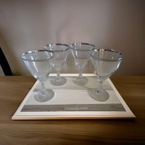 Peut inclure: Quatre verres à pied transparents avec des bords argentés sont disposés sur un plateau en bois. Les verres sont de conception classique, parfaits pour servir des cocktails. Le plateau présente un motif de grain de bois clair.