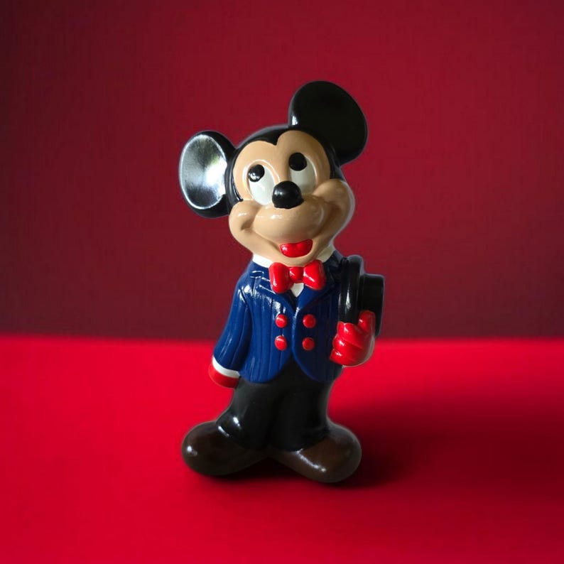 K&ouml;nnte beinhalten: Eine Keramikfigur von Mickey Mouse in einem blauen Anzug mit roten Akzenten, die einen schwarzen Zylinderhut h&auml;lt. Die Figur hat schwarze Ohren, ein beiges Gesicht und eine rote Fliege. Der Hintergrund ist ein roter Farbverlauf.