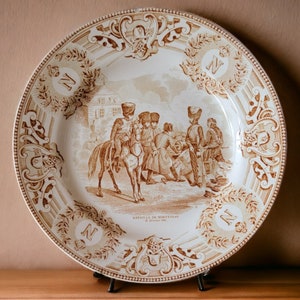 May include: A brown and white ceramic plate with a decorative border and a scene of soldiers in the center. The plate has the text "Bataille de Montreal" and "27 Janvier 1776" on it.