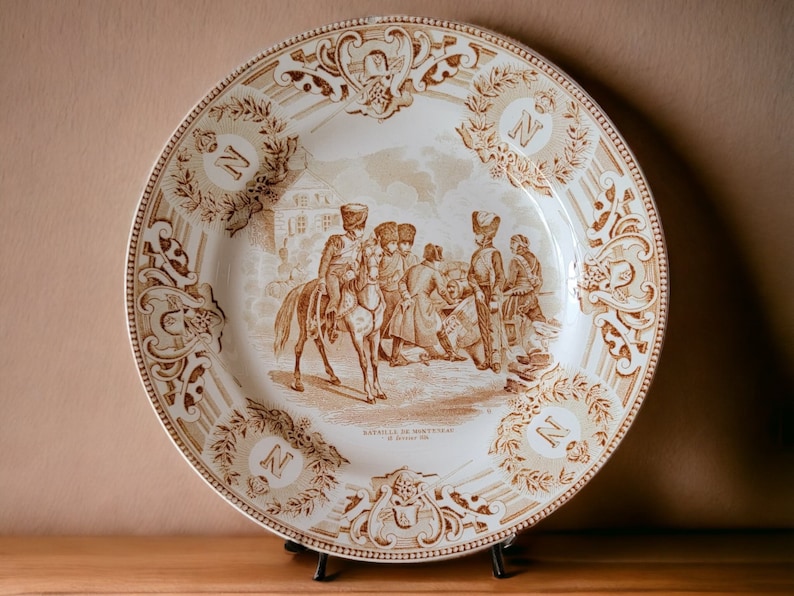 May include: A brown and white ceramic plate with a decorative border and a scene of soldiers in the center. The plate has the text "Bataille de Montreal" and "27 Janvier 1776" on it.