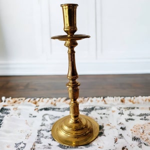 May include: A gold metal candle holder with a simple design. The holder has a round base and a tall, slender stem with a cup at the top for holding a candle.