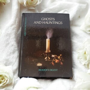 Puede incluir: Un libro negro titulado "Ghosts and Hauntings" de Reader's Digest. La portada del libro presenta una vela con humo elevándose desde ella.