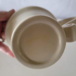 May include: A beige ceramic mug with a rounded base and a curved handle. The bottom of the mug is visible, showing a circular design. The mug appears to be clean and in good condition. The mug is likely for beverages.