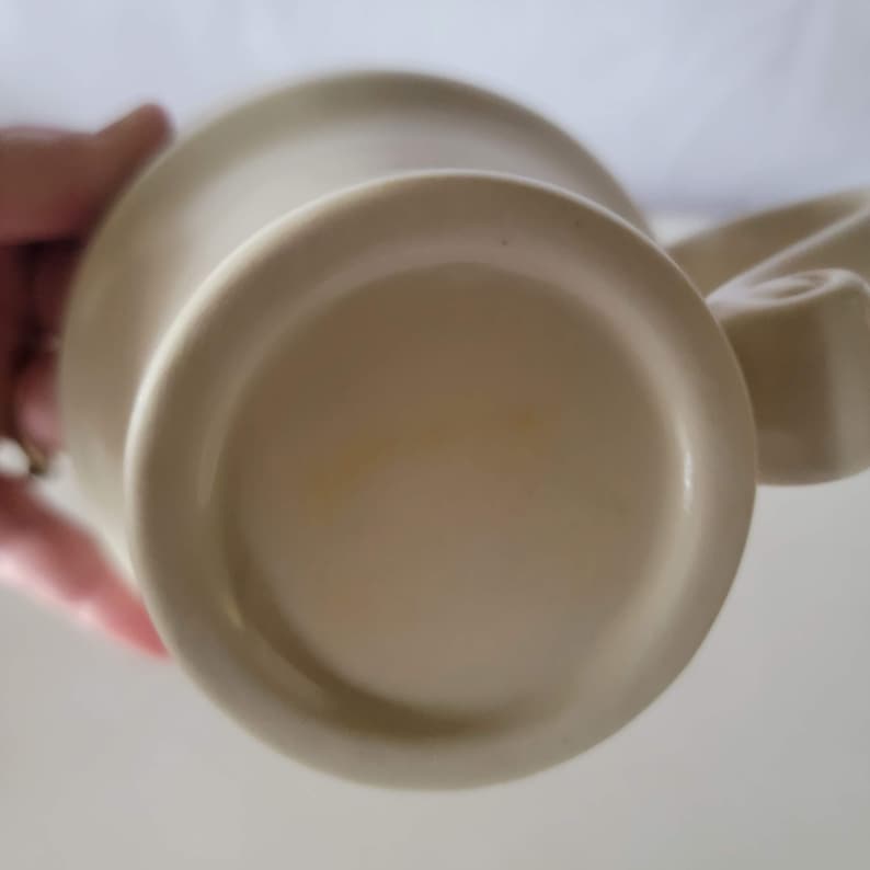 May include: A beige ceramic mug with a rounded base and a curved handle. The bottom of the mug is visible, showing a circular design. The mug appears to be clean and in good condition. The mug is likely for beverages.