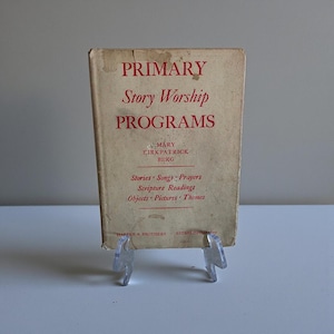 May include: A vintage book titled "PRIMARY Story Worship PROGRAMS" with red lettering on a beige cover. The book is propped up on a clear plastic stand. The book's author is Mary Kirkpatrick Berg. The book includes stories, songs, prayers, scripture readings, objects, pictures, and themes.