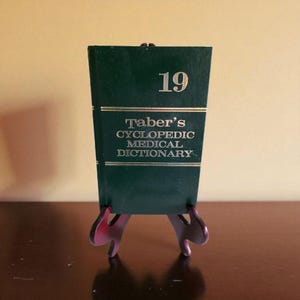 May include: A dark green book titled "Taber's Cyclopedic Medical Dictionary" with gold lettering and the number 19. The book is displayed on a small, dark red wooden stand.
