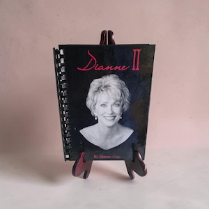 Puede incluir: Un libro encuadernado en espiral titulado "Dianne II" con un retrato de una mujer sonriente en la portada. La portada es negra con texto rosa y el libro se exhibe en un pequeño caballete de madera. El nombre del autor, Dianne Cage, está impreso en la parte inferior.