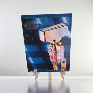 May include: A trophy with a rectangular top and a cylindrical base, displayed on a stand. The background features a blue design with stars and stripes, and the text "2002 Brickyard Four Hundred".