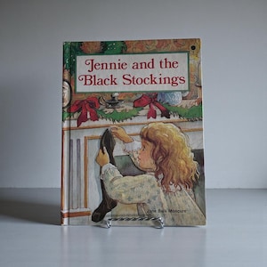 Op de afbeelding: Een vintage kinderboek getiteld "Jennie and the Black Stockings". De omslagillustratie toont een jong meisje dat een zwarte kous ophangt bij een open haard versierd met kerstversieringen. Het boek wordt vastgehouden door een metalen displaystandaard.