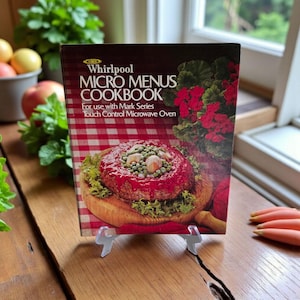 Puede incluir: Un libro de cocina vintage titulado "Whirlpool Micro Menus Cookbook" se muestra sobre una superficie de madera. La portada presenta una foto de un plato cocinado y texto. El libro está apoyado por un soporte de plástico transparente. Se ve un patrón de cuadros rojos y blancos.