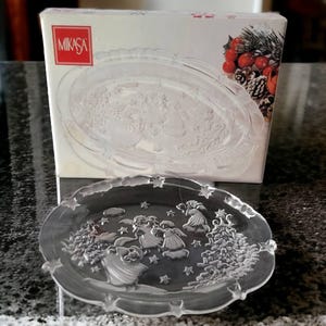 May include: A clear glass serving platter with a raised design of angels and trees. The platter is made by Mikasa and is in the shape of an oval.