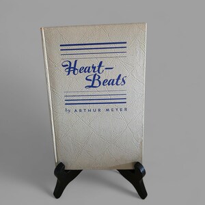 May include: A vintage book titled "Heart-Beats" by Arthur Meyer, displayed on a black easel. The book cover is a light beige with blue text and horizontal lines. The cover has a textured, dotted pattern.