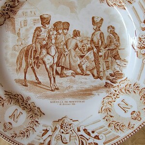 May include: A white ceramic plate with a brown design depicting a battle scene. The scene shows soldiers on horseback and on foot, with cannons and other military equipment. The text "Bataille de Monteau - 18 Fevrier 1814" is printed on the plate.