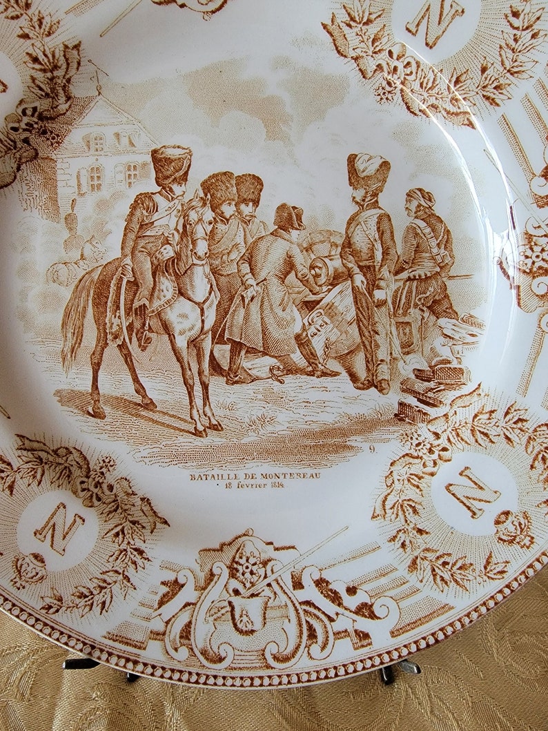 May include: A white ceramic plate with a brown design depicting a battle scene. The scene shows soldiers on horseback and on foot, with cannons and other military equipment. The text "Bataille de Monteau - 18 Fevrier 1814" is printed on the plate.