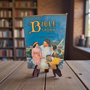 May include: A vintage book titled "The Bible Story, Volume Nine" by Arthur S. Maxwell, displayed on a wooden stand. The book cover features a blue background with a religious illustration. The stand is a dark burgundy color.
