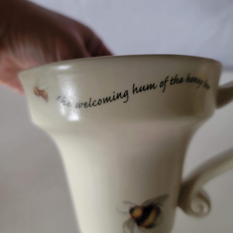May include: Cream-colored ceramic mug with a bee illustration and the text "the welcoming hum of the honey bee." The mug has a curved handle and a flared rim. A bee graphic is visible near the bottom of the mug.