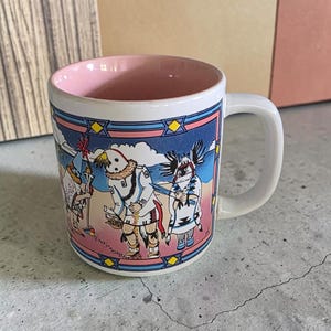 May include: A white ceramic mug with a light pink interior and a handle. The exterior features a colourful illustration of three figures in traditional attire on a beach, framed by a blue and pink border with yellow diamond accents.