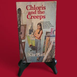 May include: A vintage paperback book titled "Chloris and the Creeps" by Kin Platt. The cover features a young girl in an orange shirt and jeans, with the title in red. The book is displayed on a black easel against a red background.