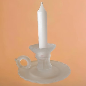 Puede incluir: Una vela blanca se encuentra en un candelero de vidrio esmerilado con un asa decorativa. El candelero tiene una base festoneada.