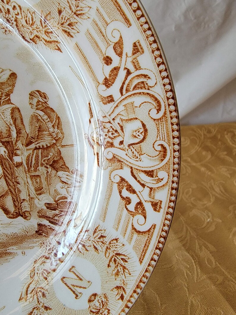 May include: A white ceramic plate with a brown floral design and a brown border. The plate has a letter "N" in the center and the number "9" on the bottom.