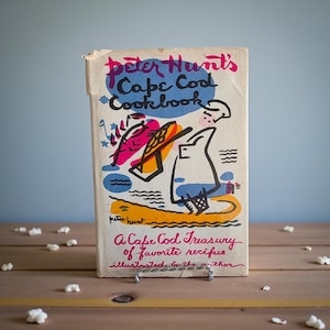 Puede incluir: Un libro de cocina vintage titulado "Peter Hunt's Cape Cod Cookbook" con una ilustración colorida. La portada presenta un diseño caprichoso con texto en rojo, azul y rosa. El libro está apoyado en un soporte metálico, con palomitas de maíz esparcidas.