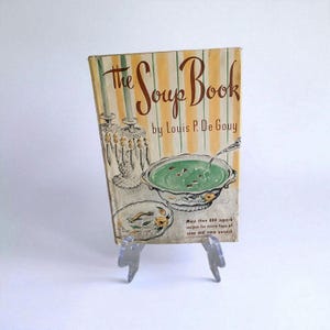 Puede incluir: Libro de cocina vintage titulado "The Soup Book" de Louis P. De Gouy. La portada presenta un fondo a rayas verdes y amarillas con ilustraciones de cuencos de sopa. El libro se exhibe sobre un soporte de plástico transparente.