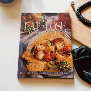 May include: A cookbook titled "Betty Crocker's New Eat and Lose Weight" with a photo of a seafood dish on the cover. The dish includes shrimp, rice, and vegetables. The text on the cover reads "Three Easy Steps to Lose Weight and Feel Great!"