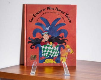 Könnte beinhalten: Ein Kinderbuch mit dem Titel "The Emperor Who Hated Yellow" wird auf einer Holzoberfläche ausgestellt. Das Cover zeigt eine Illustration eines Cartoon-Kaisers mit einem karierten Kissen und einer gelbhaarigen Figur. Das Buch wird von einem transparenten Acrylständer gehalten.