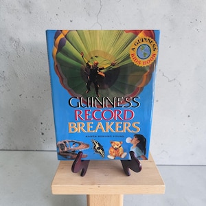 May include: A hardcover book titled Guinness Record Breakers by Karen Romano Young. The blue cover features a person skydiving with a large parachute, a dinosaur skull, a butterfly, a teddy bear, and a child blowing a bubble, with a gold seal.