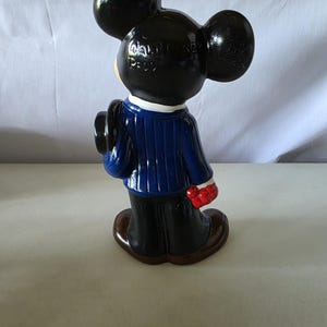 K&ouml;nnte beinhalten: Eine Keramikfigur von Mickey Mouse, von hinten gesehen, mit einem blauen Anzug, einer schwarzen Hose und braunen Schuhen. Die Figur hat schwarze Ohren und einen schwarzen Hut. Der Text "WALT DISNEY PROD" ist auf der R&uuml;ckseite des Kopfes zu sehen.