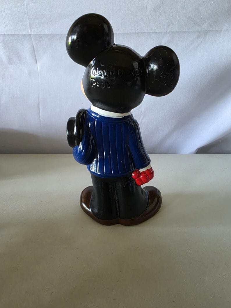 K&ouml;nnte beinhalten: Eine Keramikfigur von Mickey Mouse, von hinten gesehen, mit einem blauen Anzug, einer schwarzen Hose und braunen Schuhen. Die Figur hat schwarze Ohren und einen schwarzen Hut. Der Text "WALT DISNEY PROD" ist auf der R&uuml;ckseite des Kopfes zu sehen.