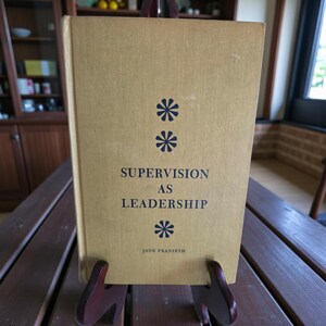 Puede incluir: Un libro de tapa dura titulado "Supervision as Leadership" con una cubierta de color amarillo claro. La cubierta presenta tres asteriscos azul oscuro y el nombre del autor, Jane Franseth, en la parte inferior. El libro se muestra en un soporte de madera.