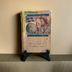 May include: Vintage book cover with a religious theme. The cover features an illustration of a man holding a flag, a globe, and the text "Occupy All Com." The book is propped up on a small black stand.