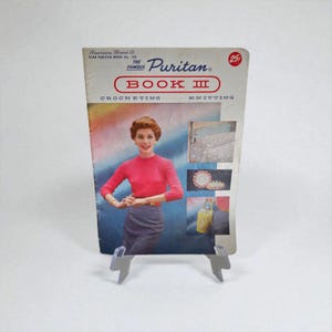 Puede incluir: Libro de patrones de ganchillo y tejido "Puritan Book III" vintage. La portada presenta a una mujer con un suéter rosa y una falda gris. El libro se muestra en un soporte de plástico transparente. El texto de la portada incluye "American Thread Co." y "Crocheting Knitting".