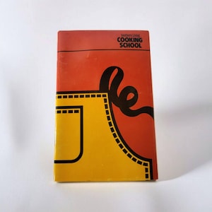 Può includere: Un libro di cucina intitolato "Southern Living COOKING SCHOOL" con un design vivace. La copertina presenta una grafica di grembiule giallo su sfondo rosso. Un design nero a forma di nastro aggiunge un tocco decorativo.