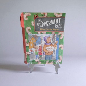 May include: A hardcover book titled "The Peppermint Race" by Dian Curtis Regan, illustrated by Anna Dewdney. The cover features a winter scene with children and houses, and a green background with candy cane designs.