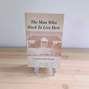 May include: A vintage book titled "The Man Who Used To Live Here" with a sepia-toned image of a house on the cover. The book is propped up on a clear plastic stand, set against a light background.