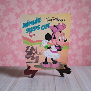 May include: A vintage book cover for "Minnie Steps Out" by Walt Disney. The cover features Minnie Mouse in a pink bow, dress, and shoes. The background has a musical note and a silhouette of a band. The book is displayed on a wooden stand.